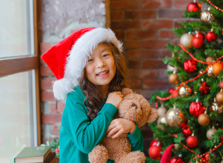 Primary aged Asian girl holding teddy bear. She's smiling with a Santa hat on, with a decorated Christmas tree behind her.