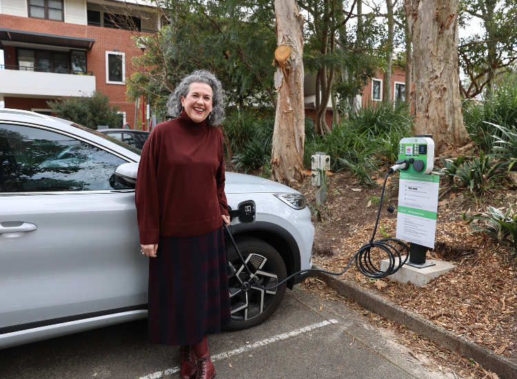 Mayor Zoë Baker standing and smiling in front of a silver car that is connected to an EV charging station with a cable.