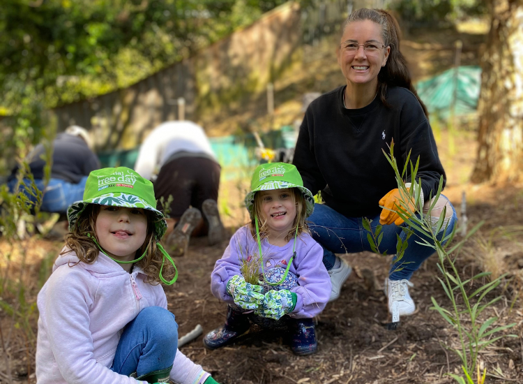 A woman and two children crouching and holding small plants, ready to plant them in the dirt.