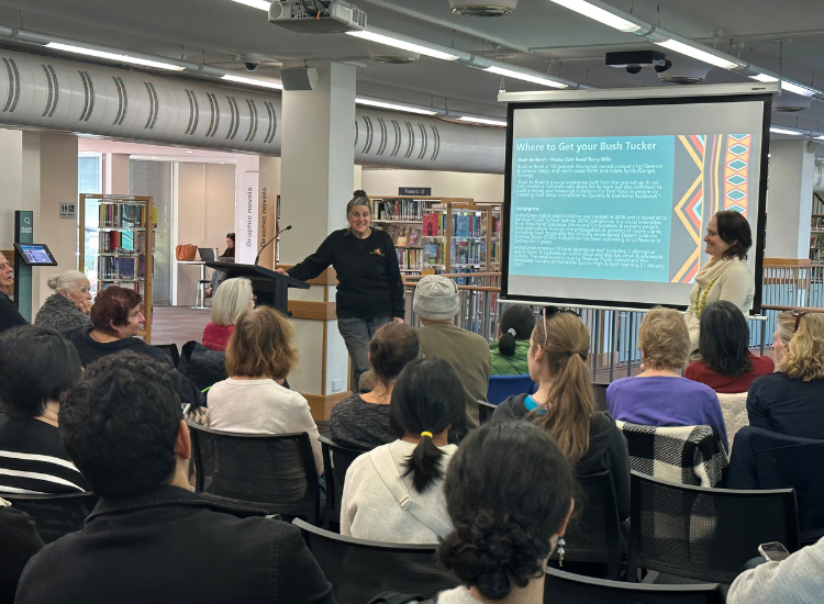 A woman standing before a small crowd beside a screen reading &#039;Where to get your bush tucker&#039;