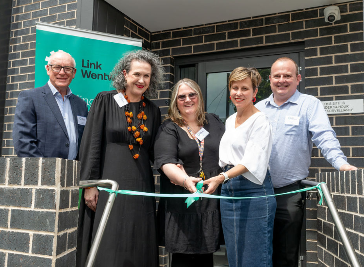 Five people smiling at the camera in front of a brick building. Two women in the middle are cutting a green ribbon with scissors
