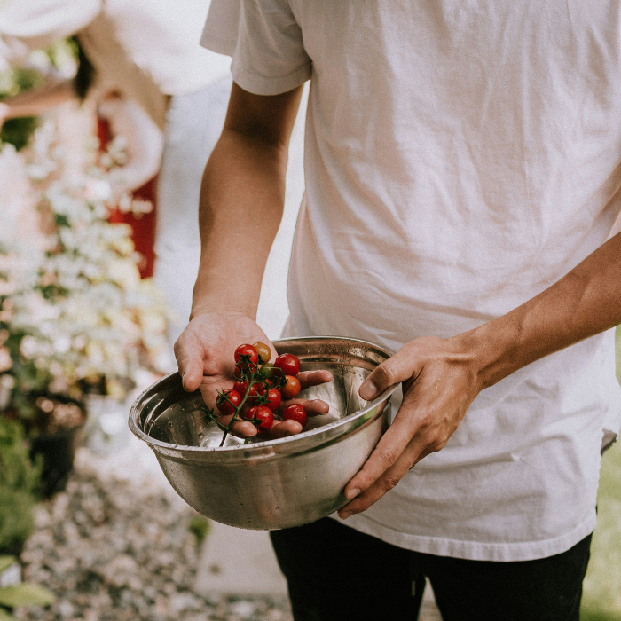 Chef standing outside near a vegetable garden, holding a metal bowl with fresh tomatoes.