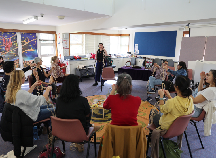 A group of women sitting on chairs in a circle, one of them standing and holding weaving string