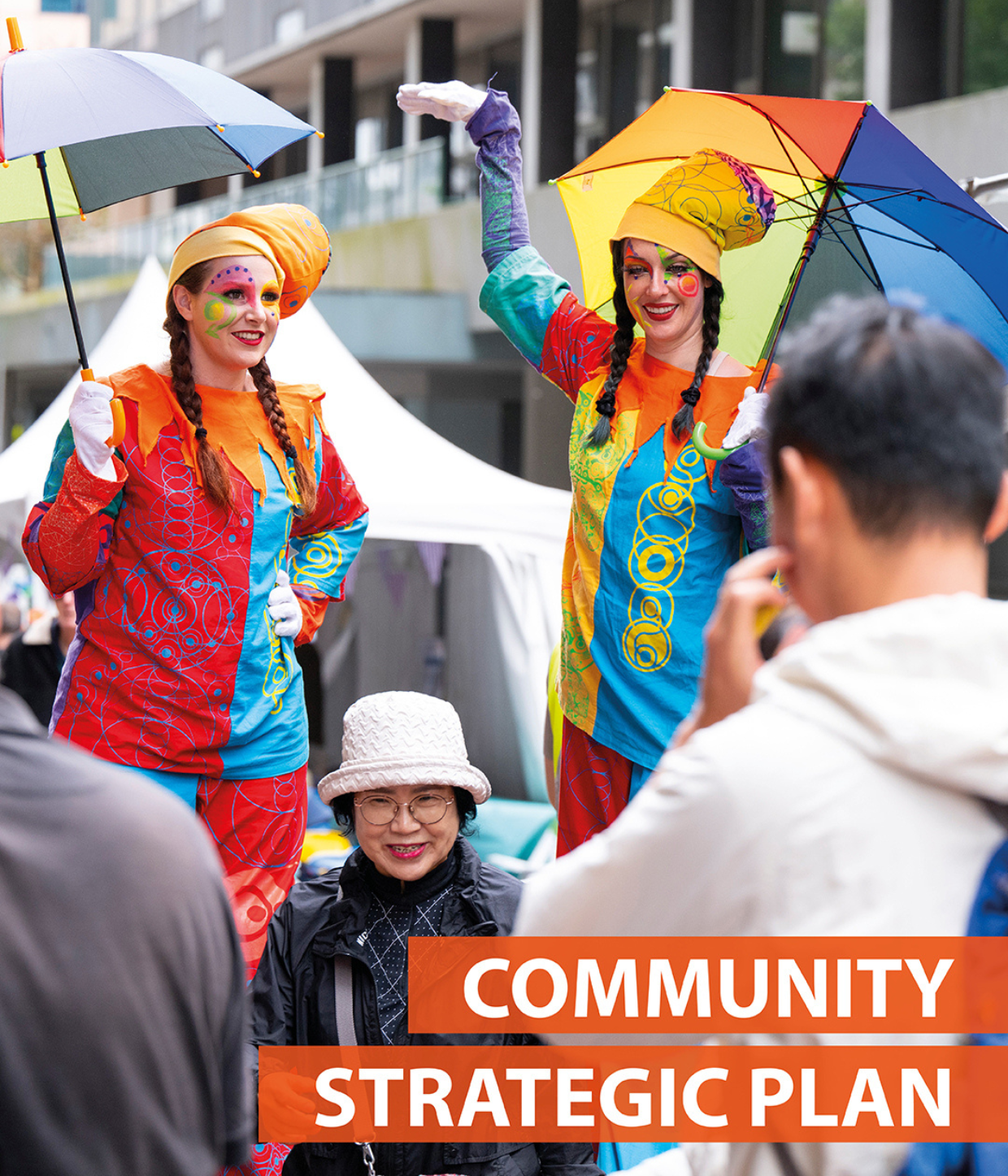 Two stilt artists with rainbow umbrella with long brown hair and theatre makeup. A woman stands in front smiling.