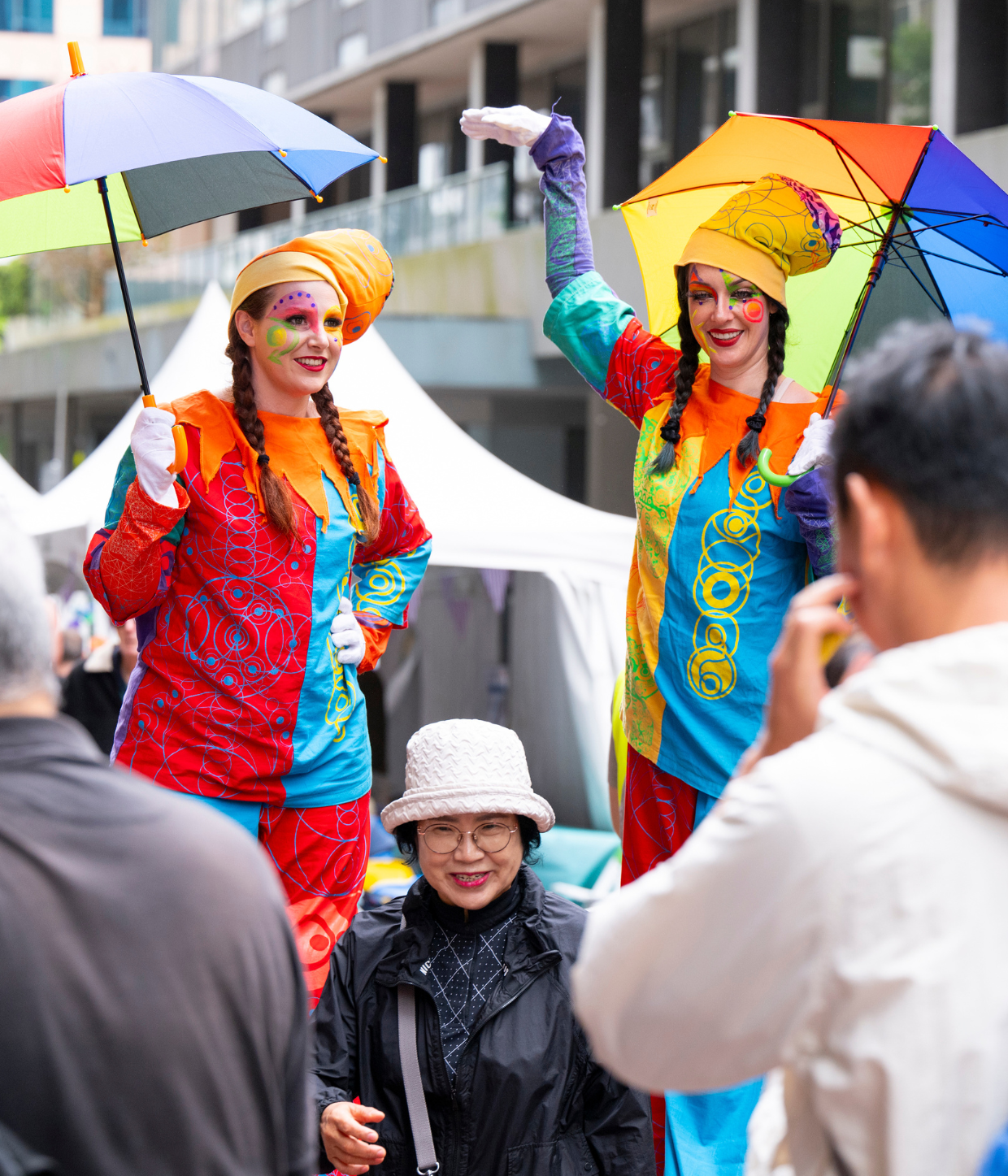 Two stilt artists with rainbow umbrella with long brown hair and theatre makeup