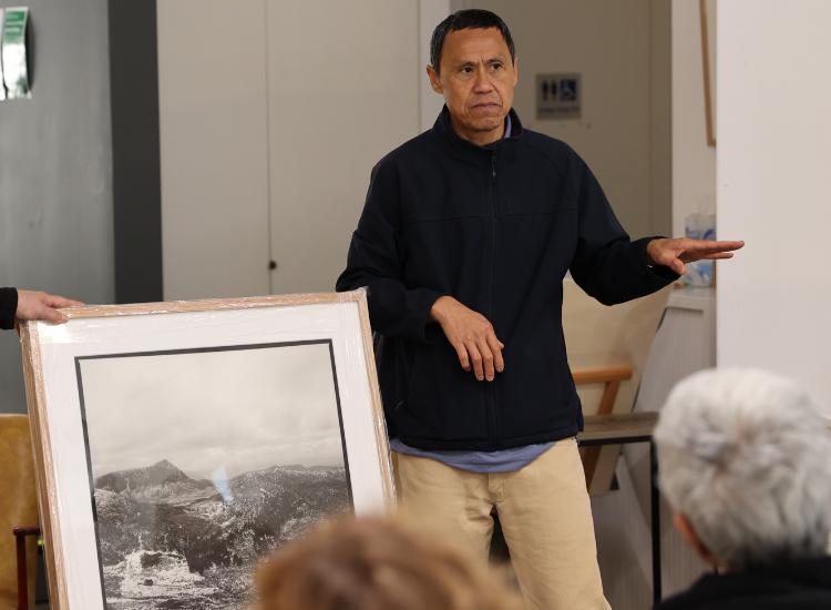 A man stands indoors speaking to a seated audience, gesturing with one hand; he is beside a large framed black-and-white landscape photograph