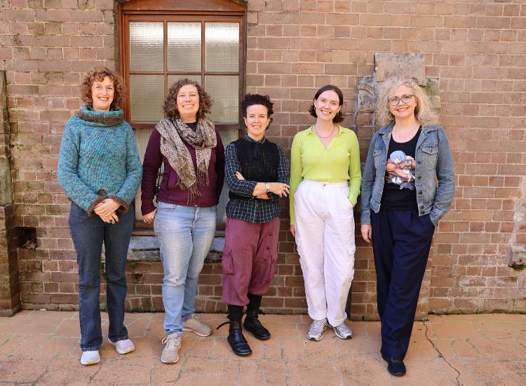 Five women stand in a row outside against a brick wall, smiling at the camera. They are dressed casually in sweaters, pants, and scarves.
