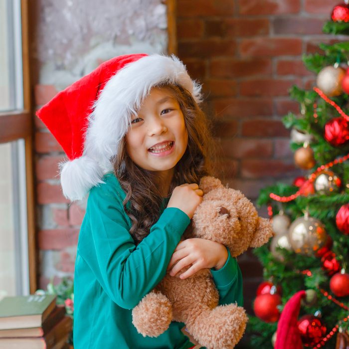 Primary aged Asian girl holding teddy bear. She's smiling with a Santa hat on, with a decorated Christmas tree behind her.