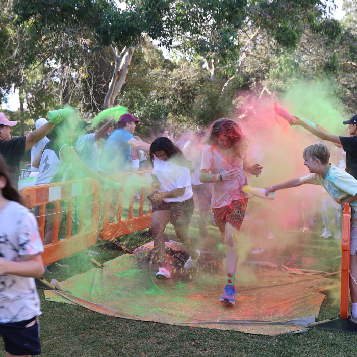 People participating in a colour run. Two young people running across a white sheet while other people throw colourful holi powder on them from behind barriers.