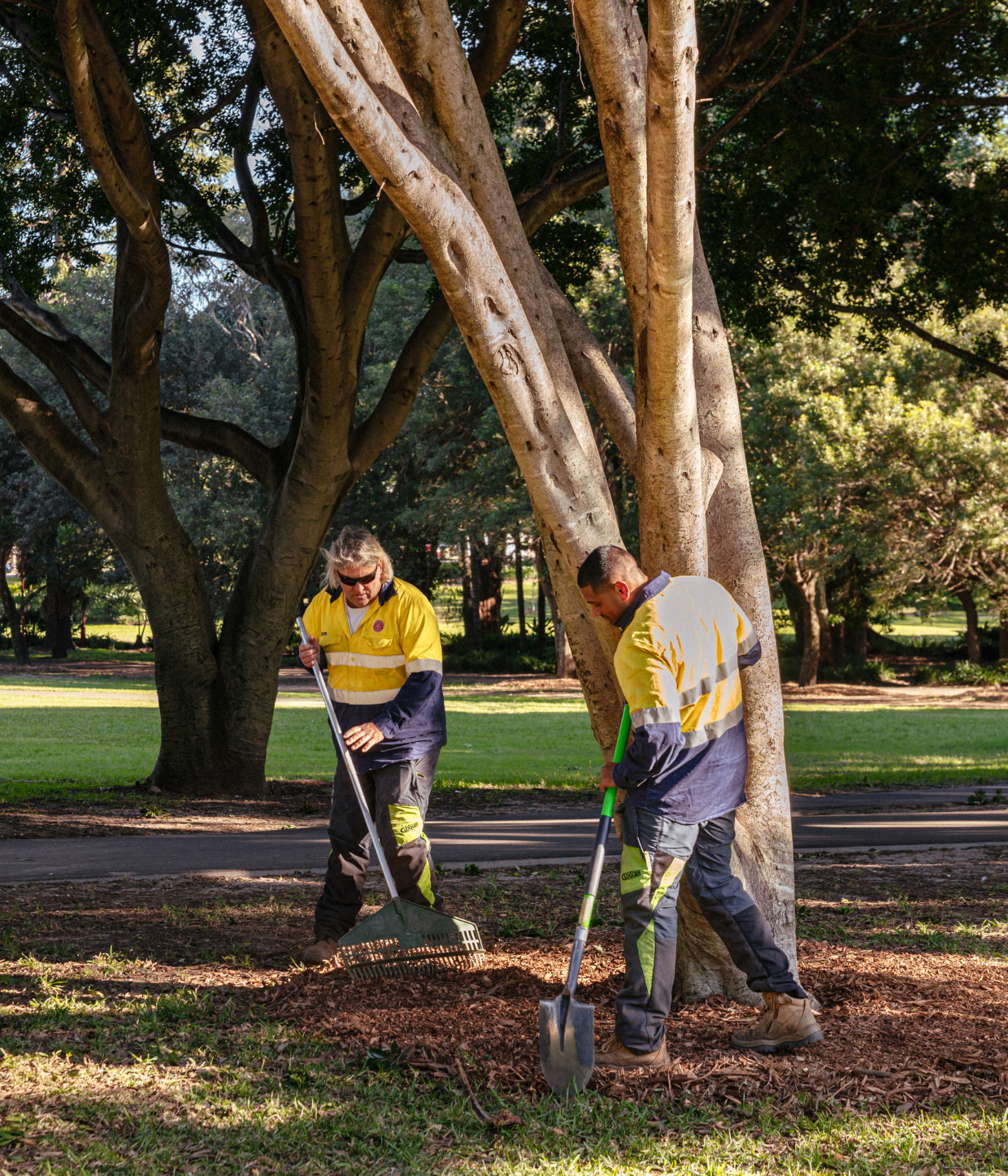 Two Council staff wearing high vis standing under a tree. One holds a rake and the other holds a spade.