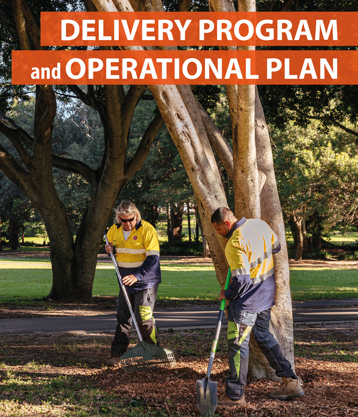 Two Council staff wearing high vis standing under a tree. One holds a rake and the other holds a spade
