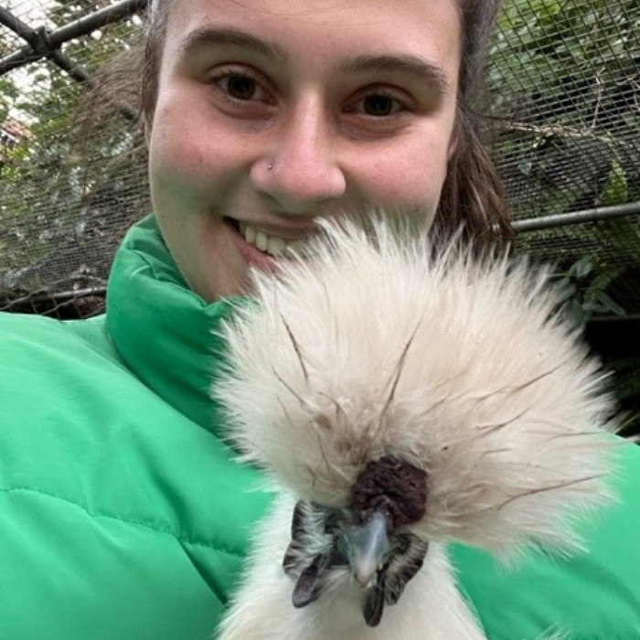 A close up shot of a woman in a green jacket smiling at the camera and a fluffy-headed chicken