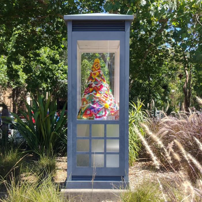 A glass phone booth houses a colorful, crocheted Christmas tree, surrounded by lush greenery and lit by bright, cheerful daylight.