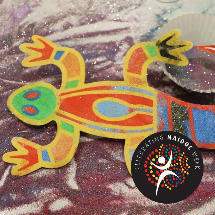 Image of a multi-coloured cut-out of an Indigenous depiction of a lizard that is lying on top of purple sand.