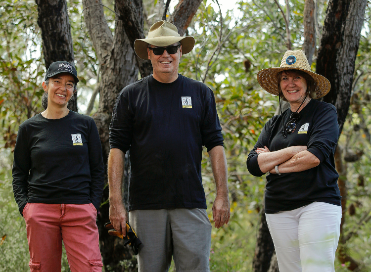 Three adults wearing hats and long-sleeve shirts stand outdoors in a bushland area, smiling at the camera.