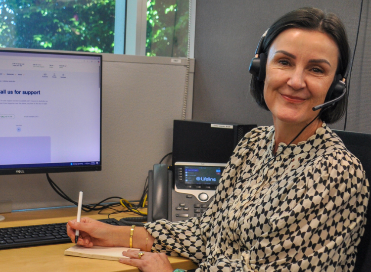 A woman sitting at a desk with a laptop, wearing a headset and smiling at the camera.
