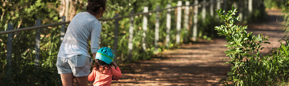 women in grey shirt holding toddler in pink shirt and green bike helmet, sitting on bike alone a dirt bike track