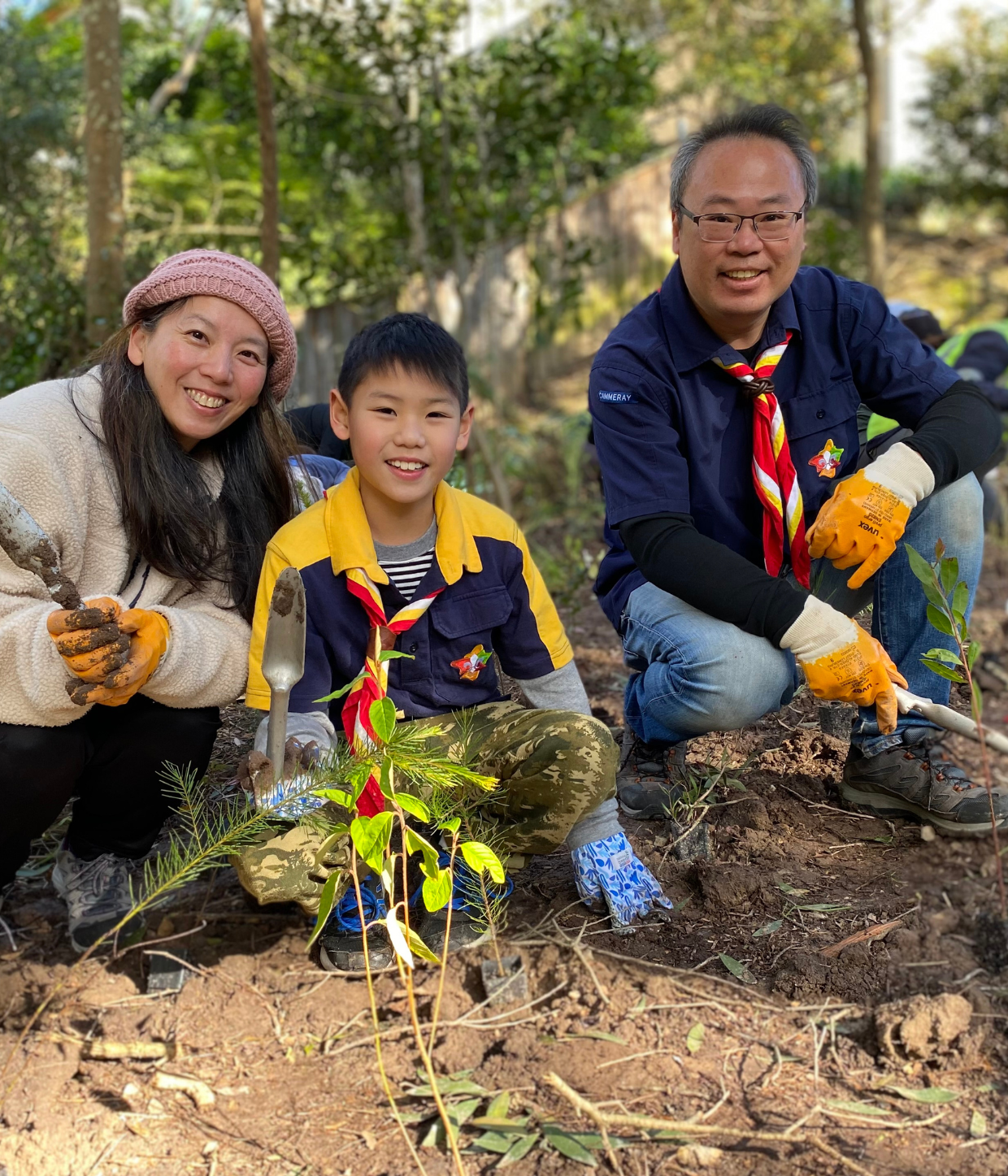 a mother, son and father crouching down smiling while holding trowels, planting a tree.