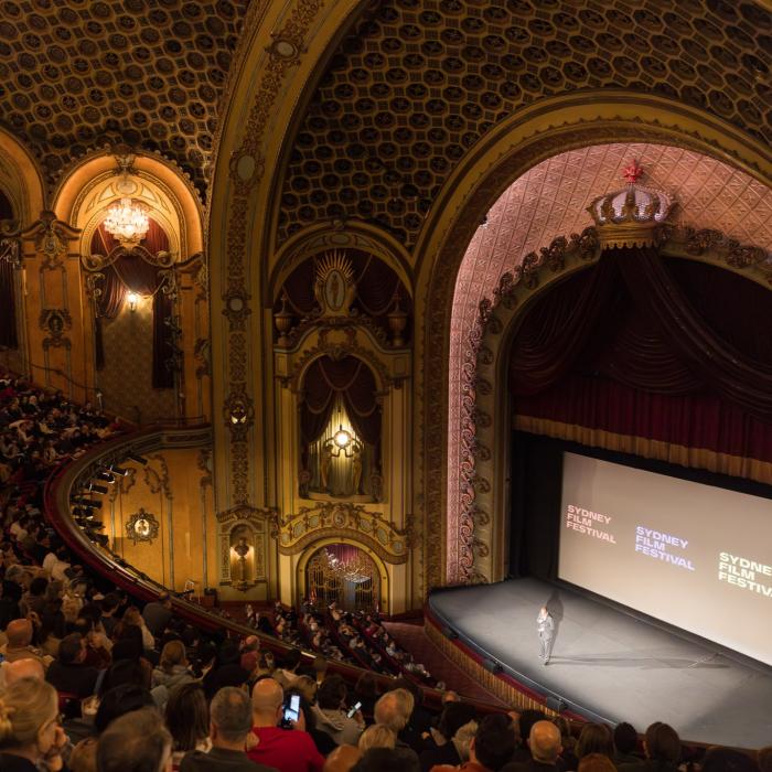 An audience faces a proscenium arch and a screen with the text 'Sydney Film Festival'