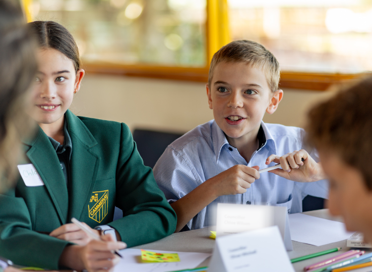 two students seated at a table holding pens to paper and smiling 
