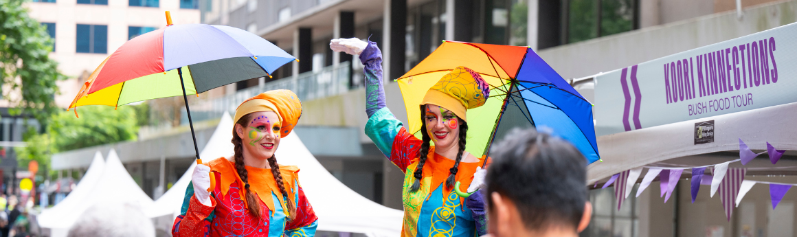 Two stilt artists with rainbow umbrella with long brown hair and theatre makeup