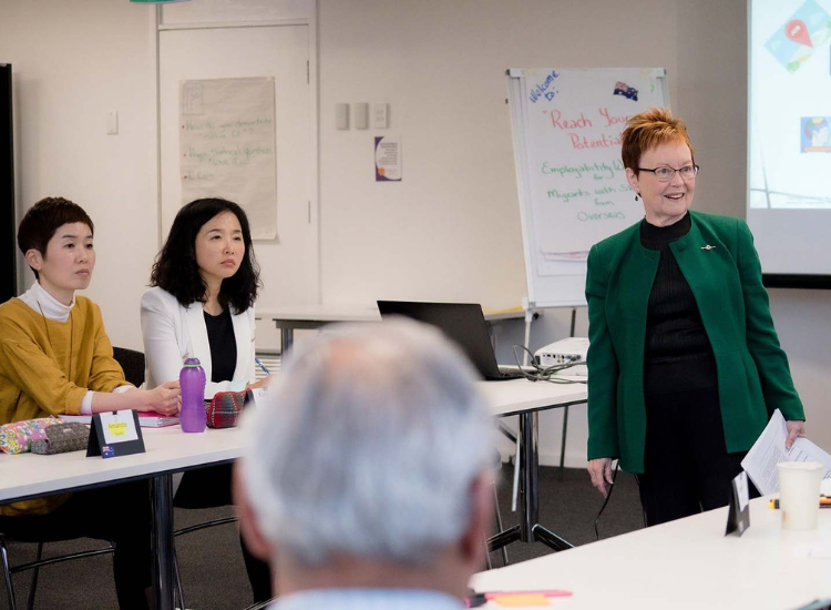 A workshop facilitator stands in front of a projector screen, smiling at workshop participants who are seated around her