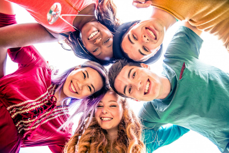 A group of five young people in brightly coloured t-shirts, standing in a circle with linked arms and smiling at the camera  below.